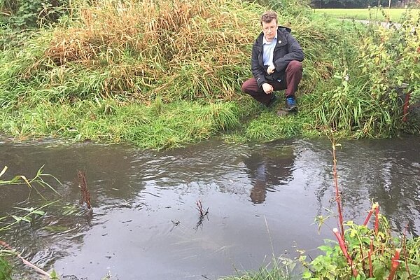 Cllr Will Frass kneeling beside a flooded Fairywell Brook